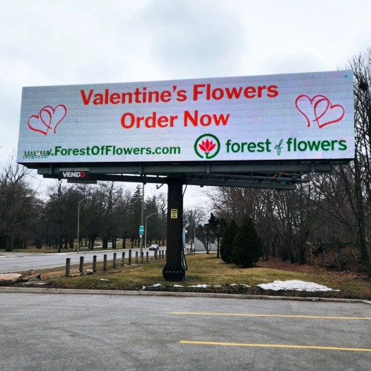 Outdoor digital billboard advertising Valentineโs Day flowers with the message โValentineโs Flowers Order Nowโ and the Forest of Flowers logo, displayed along a roadside in winter.