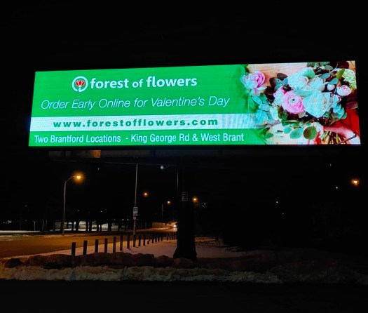 Illuminated Forest of Flowers digital billboard at night promoting Valentineโs Day flower delivery, featuring โOrder Early Online for Valentineโs Day,โ forestofflowers.com, and two Brantford florist locations on King George Road and West Brant.